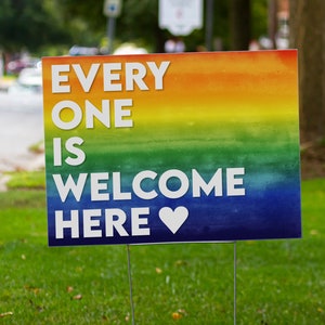 May include: A sign with a rainbow background and the text "EVERY ONE IS WELCOME HERE" with a white heart.