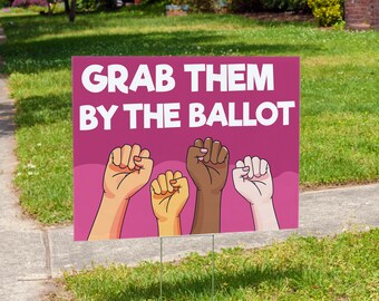 May include: Pink yard sign with the text "Grab Them By The Ballot" in white. Four raised fists of different skin tones are depicted below the text.