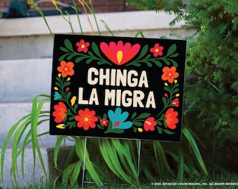 May include: A black yard sign with the text "CHINGA LA MIGRA" in white, surrounded by a floral border of red, orange, and blue flowers. The sign is displayed outdoors, set in grass.