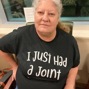 May include: A woman wearing a black t-shirt with the text "I just had a joint" printed in white. She is sitting in a chair and looking at the camera.