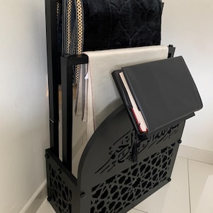 Può includere: Un porta tappeto da preghiera in legno nero con un design geometrico decorativo. Contiene diversi tappeti da preghiera e un libro. Il supporto presenta calligrafia araba e un nappa.