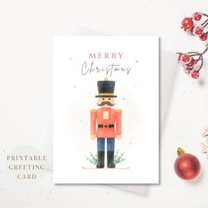 Op de afbeelding: Een witte wenskaart met een aquarelillustratie van een notenkraker-soldaat met een zwarte hoed, rood jasje en blauwe broek. De tekst MERRY Christmas staat in bruin boven de figuur gedrukt, omringd door kleine, kleurrijke sterretjes.