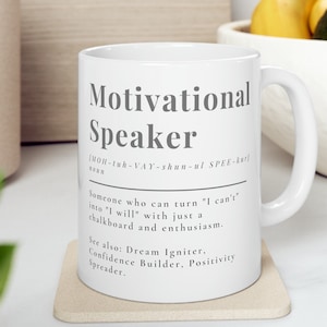 May include: White ceramic mug with a black and gray design. The mug features the word "Motivational Speaker" with a definition and pronunciation guide. The text reads: "Someone who can turn "I can't" into "I will" with just a chalkboard and enthusiasm. See also: Dream Igniter, Confidence Builder, Positivity Spreader."