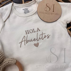 Op de afbeelding: Crème baby-romper met de geborduurde tekst "HOLA Abuelitos" en een hartje. Een houten label met "Stitched by Meg" is zichtbaar. Een gehaakte berenrammelaar is in de linker benedenhoek.