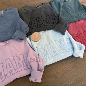 以下が含まれることがあります： 「MAMA」という単語が大きく太字で刺繍された、5色のスウェットシャツ。スウェットシャツの色は、青、グレー、ピンク、水色、赤です。