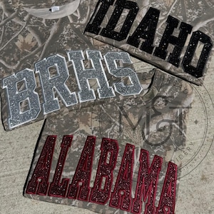 May include: Three short-sleeve t-shirts with a camouflage pattern. The top shirt features the word IDAHO in black glitter letters. The middle shirt displays BRHS in silver glitter letters. The bottom shirt shows ALABAMA in dark red glitter letters.