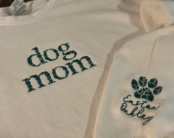 May include: White sweatshirt with green embroidered text "dog mom" on the front and a green embroidered paw print with the text "Easton Oakley" on the sleeve.