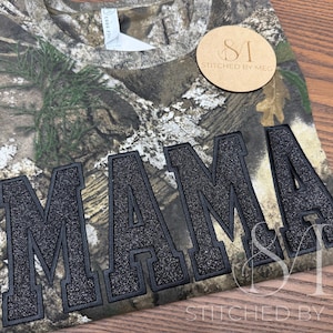 May include: A camouflage-patterned t-shirt with the word "MAMA" in large, black, glittery letters across the front. The shirt has a V-neck and a tag. A small, round, tan-colored tag with the text "STITCHED BY MEG" is also visible.