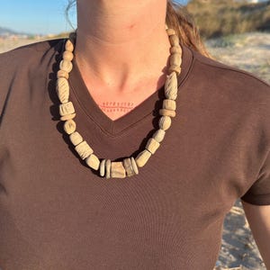 May include: A beige wooden beaded necklace with various shapes and sizes. The necklace is worn against a brown v-neck shirt. The beads have a natural, organic appearance, suggesting a handcrafted design. The necklace is a statement piece.