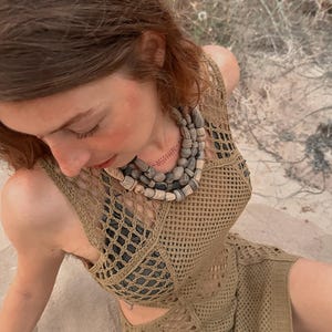 May include: A tan crochet dress with a matching top and a multi-strand beaded necklace. The dress has a net-like design with cutouts. The necklace features various shades of gray and tan beads. The outfit is worn outdoors.