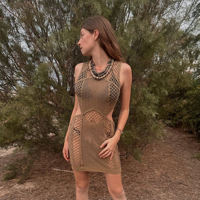 May include: A sleeveless, tan crochet dress with cut-out sides. The dress features a net-like pattern and a mini skirt length. A wooden bead necklace is worn with the dress. The background is a natural outdoor setting with green foliage.