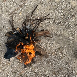 May include: A small campfire with bright orange flames burning. The fire is made of charred wood and twigs, set on a gravelly surface. A shoe is visible in the lower right corner.