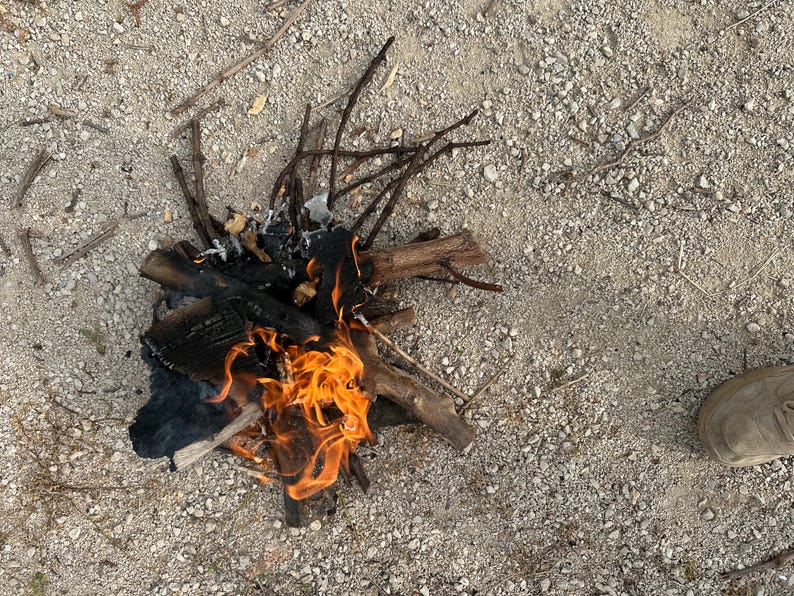 May include: A small campfire with bright orange flames burning. The fire is made of charred wood and twigs, set on a gravelly surface. A shoe is visible in the lower right corner.