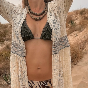 May include: A cream-colored lace kimono with intricate floral patterns is worn over a dark green bikini top and zebra-print bikini bottoms. A layered beaded necklace adds a natural touch. The scene is set outdoors with a sandy dune backdrop.