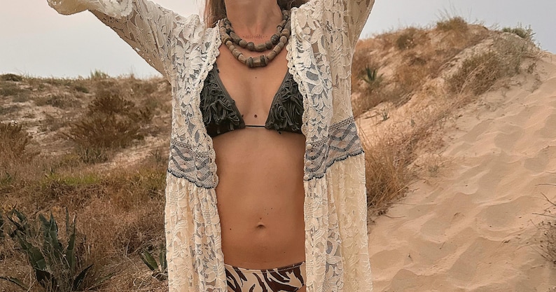 May include: A cream-colored lace kimono with intricate floral patterns is worn over a dark green bikini top and zebra-print bikini bottoms. A layered beaded necklace adds a natural touch. The scene is set outdoors with a sandy dune backdrop.