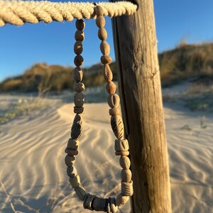 May include: A handcrafted wooden bead necklace with various shapes and textures hangs from a rope. The necklace is displayed against a backdrop of a wooden post, sand, and a clear blue sky, suggesting a beach or coastal theme.