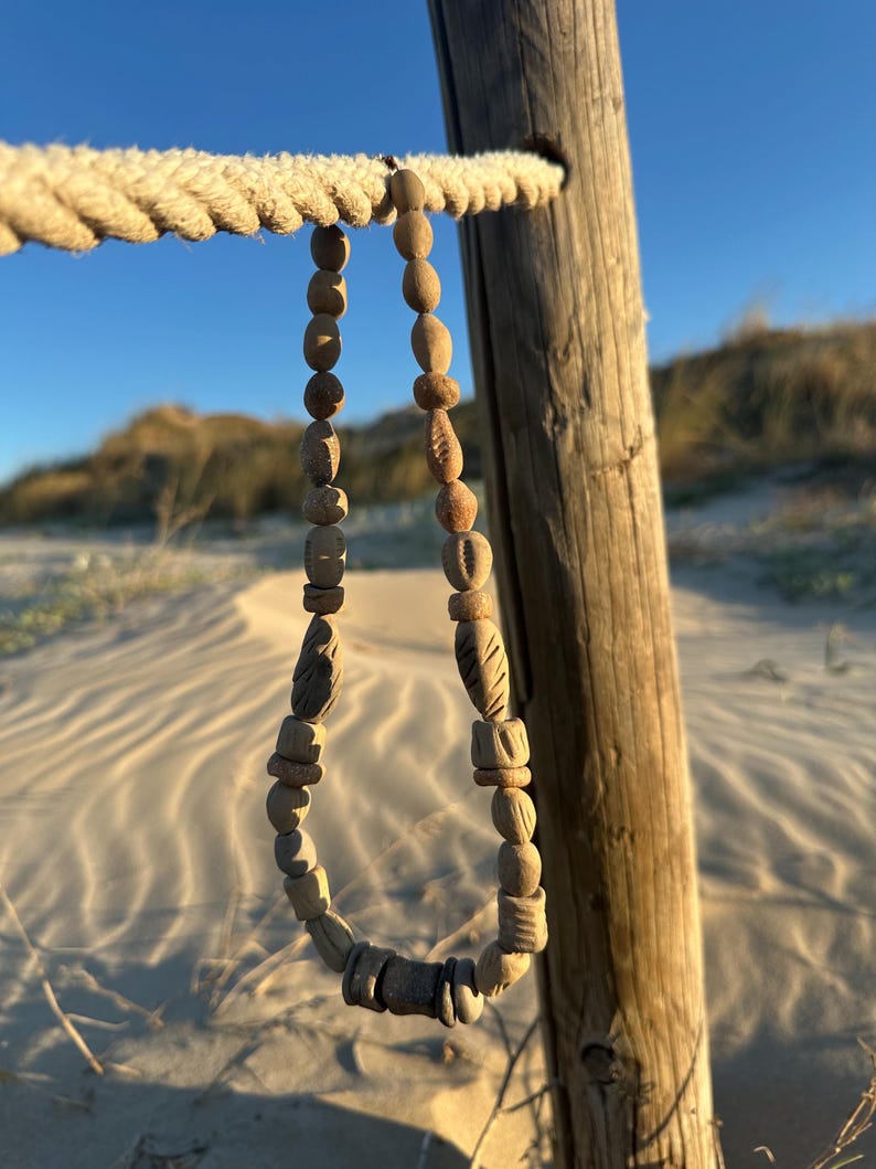 May include: A handcrafted wooden bead necklace with various shapes and textures hangs from a rope. The necklace is displayed against a backdrop of a wooden post, sand, and a clear blue sky, suggesting a beach or coastal theme.