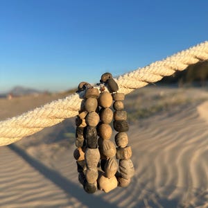 May include: A handmade bracelet composed of various wooden beads in shades of brown and gray. The bracelet is hanging on a thick, off-white rope, with a sandy beach and blue sky in the background. The beads have unique shapes and textures.