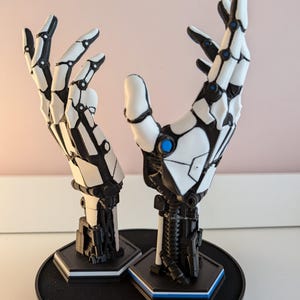 Puede incluir: Dos manos robóticas blancas con detalles negros, posadas con las palmas abiertas, se muestran en una base circular negra con plataformas hexagonales.