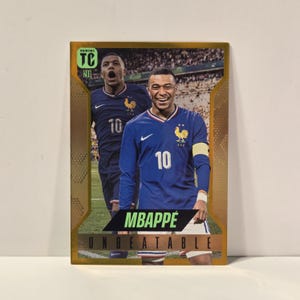 Può includere: Una figurina rettangolare Panini TC con bordo dorato e una fotografia di Kylian Mbappé con la maglia da calcio blu della Francia con il numero 10. La carta mostra il testo MBAPPÉ e UNBEATABLE in grassetto su uno sfondo raffigurante uno stadio.