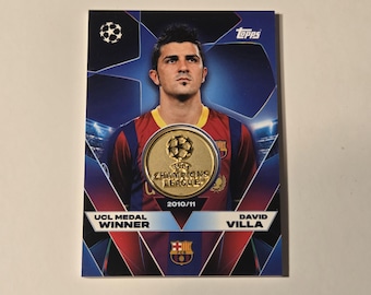 May include: Topps trading card featuring David Villa, a UCL Medal Winner. The card has a gold medallion with "Champions League 2010/11" text. The background is blue with a soccer ball and stadium lights.