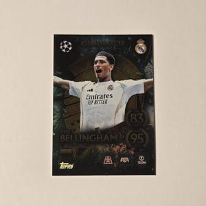 Op de afbeelding: Een voetbal trading card met Jude Bellingham in een wit shirt met de tekst "Emirates Fly Better". De kaart heeft een gouden en zwarte achtergrond met de tekst "Gladiator", "83" en "95". Het Topps logo is zichtbaar in de linker benedenhoek.