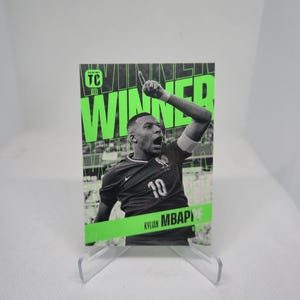 Op de afbeelding: Een Panini-verzamelkaart met Kylian Mbappé in een zwart shirt met nummer 10. De kaart heeft het woord "WINNER" in grote groene letters en de naam van de speler onderaan. De kaart wordt gepresenteerd op een transparante plastic standaard.