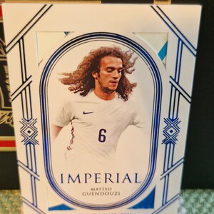 May include: A white and blue sports trading card featuring a photo of a football player wearing a white shirt with a blue stripe and the number 6. The card has the text "Imperial", "Matteo Guendouzi" and "Game Used Memorabilia".