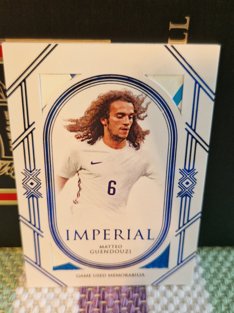 May include: A white and blue sports trading card featuring a photo of a football player wearing a white shirt with a blue stripe and the number 6. The card has the text "Imperial", "Matteo Guendouzi" and "Game Used Memorabilia".