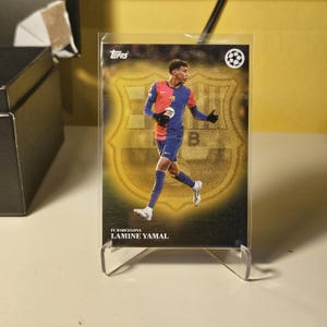 Könnte beinhalten: Eine Topps-Sammelkarte mit Lamine Yamal vom FC Barcelona. Die Karte hat einen goldenen Hintergrund mit dem Teamlogo. Yamal trägt ein rot-blaues und marinefarbenes Trikot. Die Karte befindet sich in einer Schutzhülle und wird auf einem transparenten Ständer präsentiert.