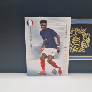 Può includere: Una carta di calcio bianca e blu con un calciatore che indossa una maglia blu con il numero 8. La carta ha un bordo dorato e il testo "MIDFIELDER" sul lato destro. Il giocatore è Aurélien Tchouameni, un calciatore francese.