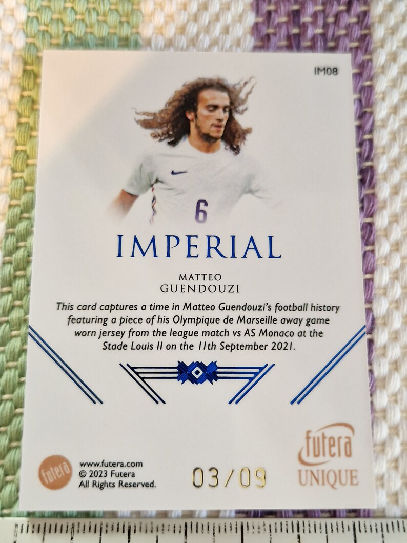 May include: A white and blue football jersey with the number 6 on the front. The jersey is worn by a player with long brown hair. The card is part of a set called "Imperial" and is numbered 03/09.