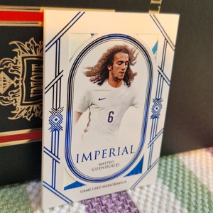 May include: A white and blue sports trading card featuring a photo of a football player wearing a white jersey with a blue number 6. The card has the text "Imperial Matteo Guendouzi Game Used Memorabilia".