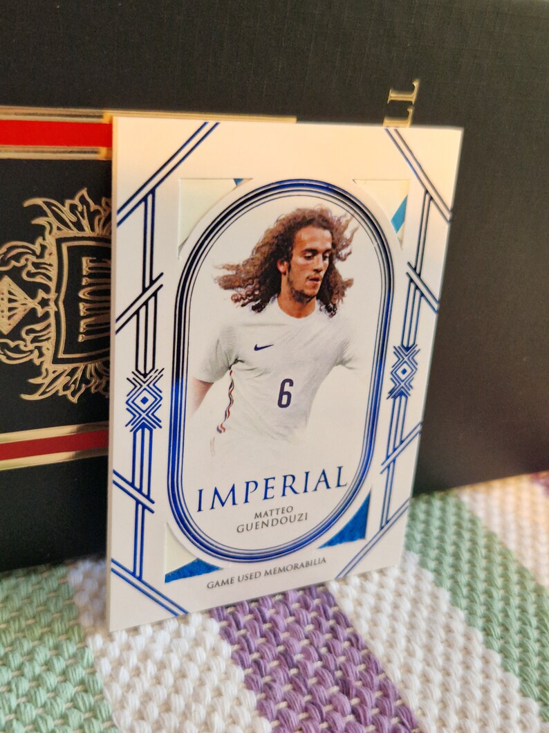 May include: A white and blue sports trading card featuring a photo of a football player wearing a white jersey with a blue number 6. The card has the text "Imperial Matteo Guendouzi Game Used Memorabilia".