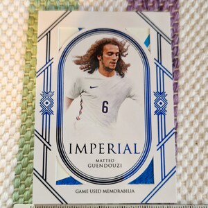 May include: A white and blue sports trading card featuring a photo of a football player wearing a white jersey with a blue number 6. The card has the text "IMPERIAL" and "MATTEO GUENDOUZI" in black text. The card also has the text "GAME USED MEMORABILIA" in black text.