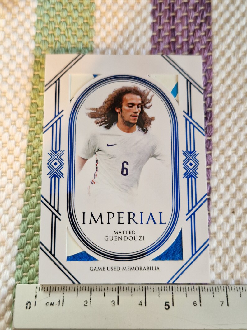 May include: A white and blue sports trading card featuring a photo of a football player wearing a white jersey with a blue number 6. The card has the text "IMPERIAL" and "MATTEO GUENDOUZI" in black text. The card also has the text "GAME USED MEMORABILIA" in black text.