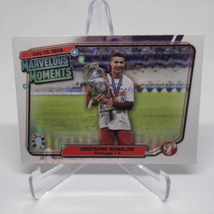 Può includere: Una carta da collezione con Cristiano Ronaldo che tiene in alto il trofeo del Campionato Europeo. La carta è della serie "Marvelous Moments" ed è datata 10 luglio 2016. La carta ha una finitura olografica e presenta uno schema di colori rosso, bianco e blu.