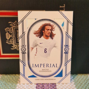 May include: A white and blue sports trading card featuring a photo of a football player wearing a white shirt with a blue number 6. The card has the text "Imperial" and "Matteo Guendouzi" in blue lettering. The card also has the text "Game Used Memorabilia" in blue lettering.