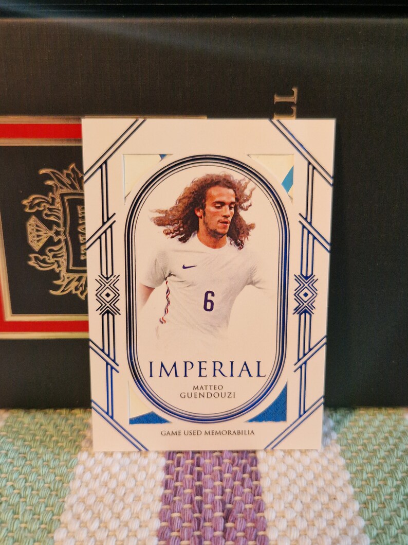 May include: A white and blue sports trading card featuring a photo of a football player wearing a white shirt with a blue number 6. The card has the text "Imperial" and "Matteo Guendouzi" in blue lettering. The card also has the text "Game Used Memorabilia" in blue lettering.