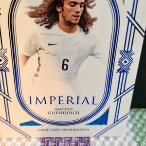 May include: A white and blue sports card featuring a photo of a football player wearing a white shirt with a blue number 6. The card has the text "Imperial" and "Matteo Guendouzi" in blue lettering. The card also has the text "Game Used Memorabilia" in blue lettering.