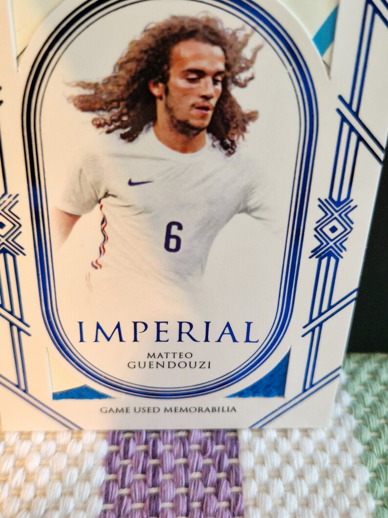 May include: A white and blue sports card featuring a photo of a football player wearing a white shirt with a blue number 6. The card has the text "Imperial" and "Matteo Guendouzi" in blue lettering. The card also has the text "Game Used Memorabilia" in blue lettering.