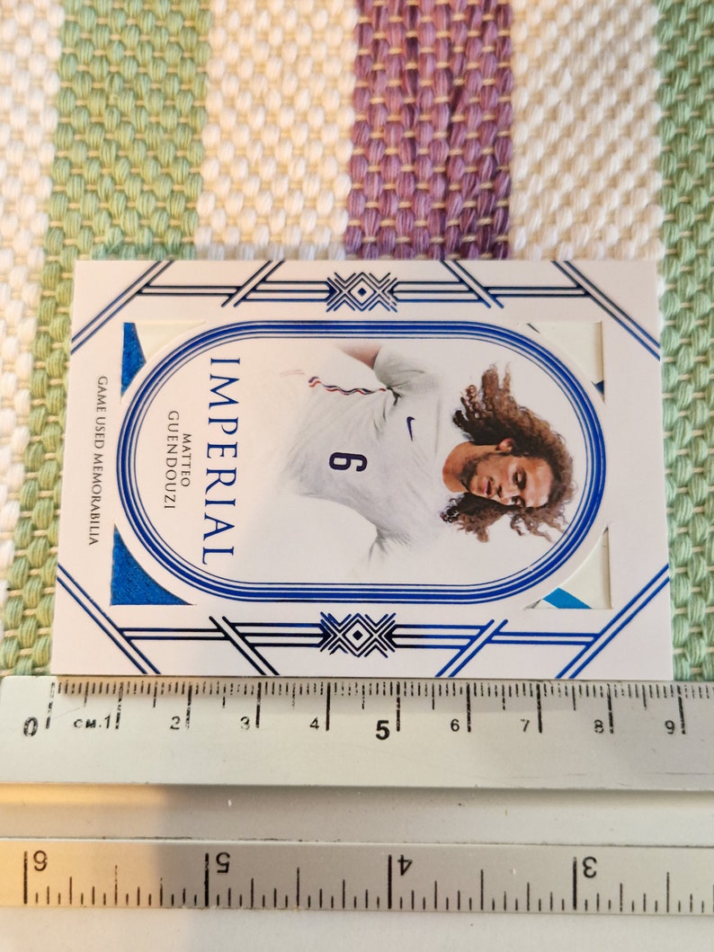 May include: A white and blue sports card featuring a photo of a football player wearing a white jersey with a blue number 6. The card has the text "IMPERIAL" and "MATTEO GUENDOUZI" on it. The card also has the text "GAME USED MEMORABILIA" on it.