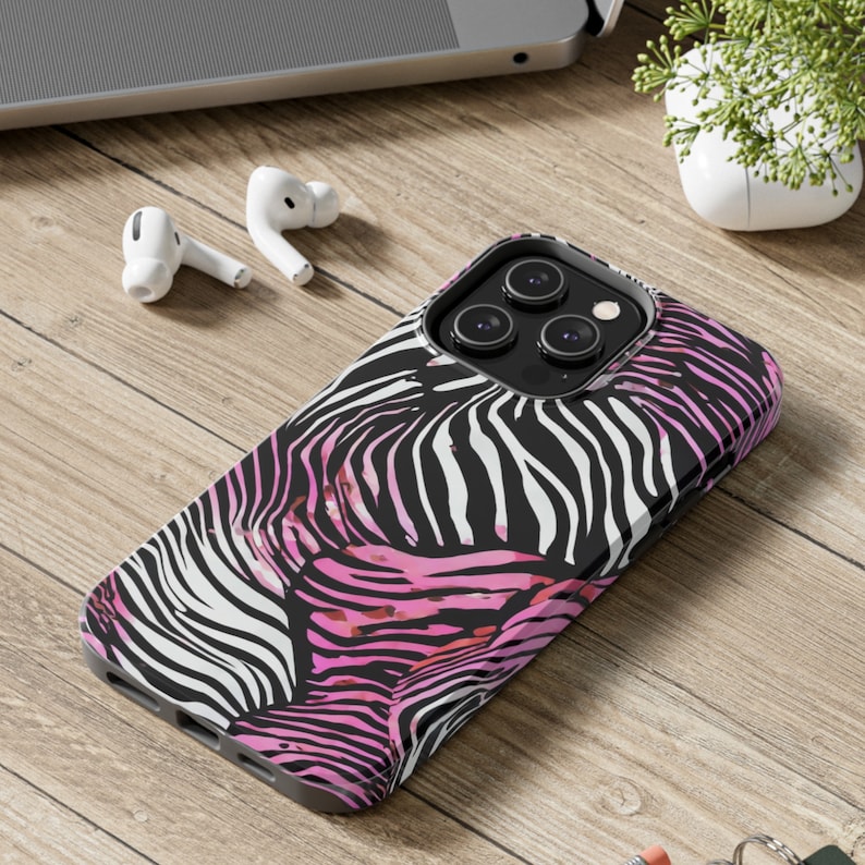 K&ouml;nnte beinhalten: Eine Handyh&uuml;lle mit einem auff&auml;lligen Zebra-Print-Design in Schwarz, Wei&szlig; und Pink. Die H&uuml;lle liegt auf einer Holzoberfl&auml;che, im Hintergrund sind wei&szlig;e Ohrh&ouml;rer und ein Laptop zu sehen. Die H&uuml;lle ist f&uuml;r ein Smartphone konzipiert.