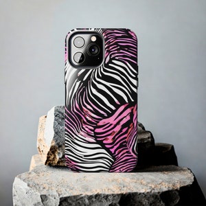 K&ouml;nnte beinhalten: Eine Smartphone-H&uuml;lle mit einem Zebra-Streifenmuster in Schwarz, Wei&szlig; und Pink. Die H&uuml;lle hat eine gl&auml;nzende Oberfl&auml;che und einen Kameraausschnitt. Die Handyh&uuml;lle wird auf einer strukturierten Steinoberfl&auml;che pr&auml;sentiert.