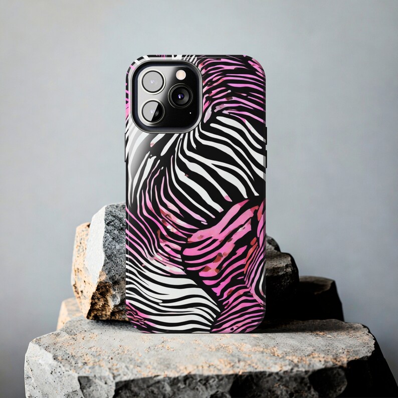 K&ouml;nnte beinhalten: Eine Smartphone-H&uuml;lle mit einem Zebra-Streifenmuster in Schwarz, Wei&szlig; und Pink. Die H&uuml;lle hat eine gl&auml;nzende Oberfl&auml;che und einen Kameraausschnitt. Die Handyh&uuml;lle wird auf einer strukturierten Steinoberfl&auml;che pr&auml;sentiert.