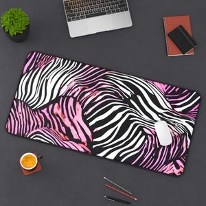 May include: A rectangular mouse pad with a zebra stripe pattern in black, white, and pink. A white computer mouse sits on the pad. A laptop, pencils, a notebook, and a small plant are also visible.