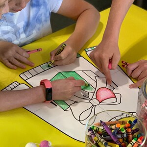May include: Two children color a drawing of a sweater with a bow and a heart on a yellow table. They are using crayons to color the drawing. There is a jar of crayons on the table.