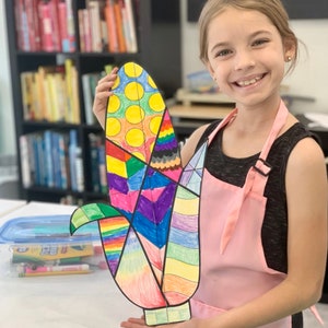 May include: A child holds up a colorful paper craft of an ear of corn. The craft is made of different colored paper squares and circles. The child is wearing a pink apron and a black tank top. The background is a bookshelf with many books.