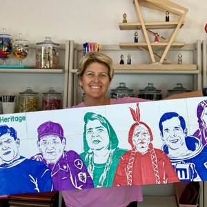 May include: A woman holds a large poster with the words "American Heritage" at the top. The poster features eight colorful portraits of people from different backgrounds, including a firefighter, a Native American, and an astronaut.