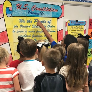 Può includere: Un gruppo di bambini si raduna intorno a un poster giallo e rosso con il testo "U.S. Constitution Preamble" e la prima riga del preambolo, "We the People of the United States, in Order..."
